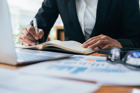 Person writing in notebook with graphs and financial metric sheets on the desk